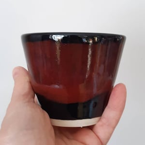 May include: A small, hand-thrown ceramic cup with a black rim and a red and black glaze.