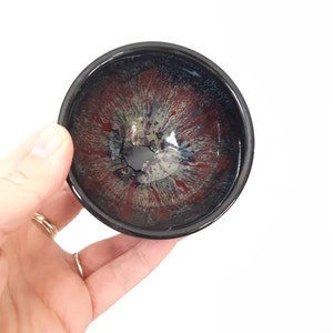 May include: A small black ceramic bowl with a red, white and blue glaze. The bowl has a crack in the glaze.