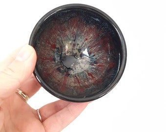 May include: A small black ceramic bowl with a red, white, and blue glaze. The bowl has a crack in the glaze.
