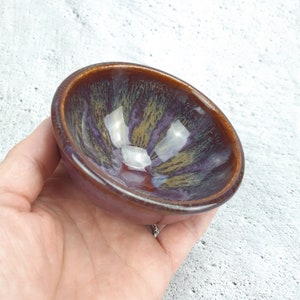 May include: A small, round ceramic bowl with a brown, purple and gold glaze. The bowl has a glossy finish and is ideal for serving small snacks or dips.