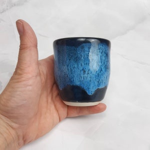 May include: A small, blue and white ceramic cup with a speckled glaze. The cup is slightly rounded and has a white rim.
