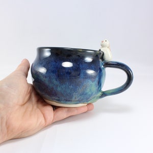 May include: A blue and green ceramic mug with a white rabbit figurine on the rim. The mug has a rounded body and a handle.
