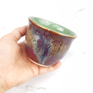 May include: A small, handmade ceramic bowl with a red, brown, and gold glaze. The inside of the bowl is glazed in a light green color.
