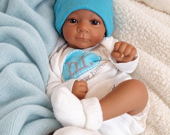 May include: A baby doll with dark skin wearing a bright blue beanie and a white long-sleeved onesie. The onesie features a blue cloud graphic with the text "hi." and "new here" printed on the front. The doll is wearing white fuzzy socks.