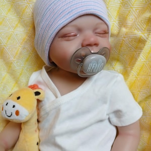 May include: A lifelike baby doll wearing a light blue and pink striped beanie, a white wrap-style onesie, and a gray pacifier with the word "HELLO". The doll is holding a yellow giraffe plush toy with orange accents.