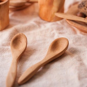 May include: Two wooden spoons on a white linen tablecloth. The spoons are made of natural wood and have a smooth, rounded shape.