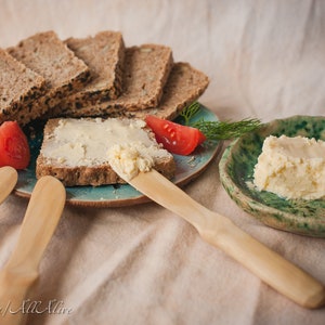 May include: A close-up of a plate with a slice of bread topped with butter, two red tomatoes, and a sprig of dill. There are four wooden butter knives and a plate with a block of butter on the side.