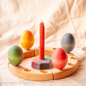 Może przedstawiać: Drewniany stojak na jajka z pięcioma gniazdami, z których każde zawiera kolorowe jajko. Czerwona świeca pali się w środku stojaka.