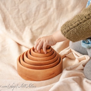 Puede incluir: La mano de un niño alcanza un juguete de apilar de madera natural. El juguete está hecho de siete anillos encajables, cada uno de un tamaño ligeramente diferente. Los anillos están apilados en forma de pirámide. El juguete está sobre una manta beige claro.