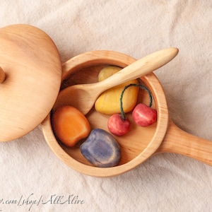 Könnte beinhalten: Ein Holztopf mit Deckel und einem Holzlöffel im Inneren. Der Topf enthält mehrere Holzfrüchte, darunter eine gelbe Birne, ein orangefarbener Pfirsich, eine violette Pflaume und zwei rote Kirschen.