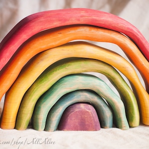 Puede incluir: Un juguete de arco de madera de colores arcoíris con siete piezas, cada una de un color diferente, apiladas una encima de la otra. Los colores son rojo, naranja, amarillo, verde, azul, añil y violeta. El juguete está hecho de madera natural y tiene un acabado liso y pulido.