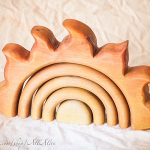 Peut inclure: Un jouet d'empilage en bois en forme d'arc-en-ciel avec une base en forme de soleil. Le jouet est fait de bois naturel et a une finition chaude de couleur miel. L'arc-en-ciel est composé de cinq arches qui s'emboîtent les unes dans les autres.