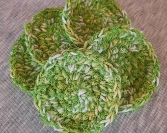 May include: A collection of handmade, round crochet scrubbies in shades of green and white. The scrubbies are arranged in a slightly overlapping pattern, showcasing their textured, looped design. The overall aesthetic is rustic and eco-friendly.