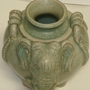 Puede incluir: Un jarr&oacute;n de cer&aacute;mica de color verde claro con acabado de esmalte craquelado, que presenta una cabeza de elefante esculpida en la parte frontal y asas decorativas en forma de cuerda en los laterales. El jarr&oacute;n tiene un cuerpo ancho y redondeado.
