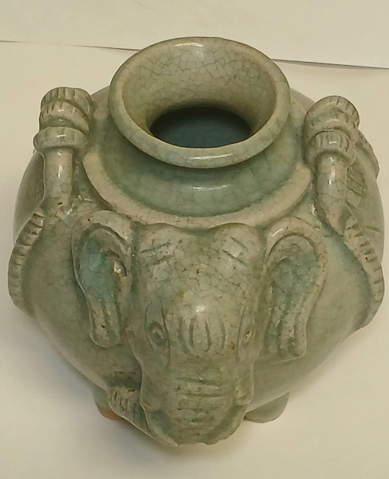 Puede incluir: Un jarr&oacute;n de cer&aacute;mica de color verde claro con acabado de esmalte craquelado, que presenta una cabeza de elefante esculpida en la parte frontal y asas decorativas en forma de cuerda en los laterales. El jarr&oacute;n tiene un cuerpo ancho y redondeado.
