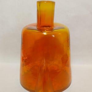 May include: A translucent orange glass bottle with a cylindrical neck and a wide, rounded body. The base features a distinct, pointed triangular protrusion extending from the center, creating a unique geometric shape in the vibrant glass.