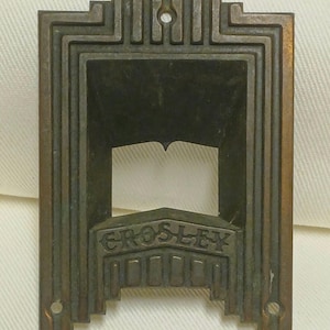 Puede incluir: Una placa protectora rectangular de metal color bronce con un borde geométrico escalonado y un recorte central. La palabra CROSLEY está grabada en letras mayúsculas sobre una serie de barras verticales en el borde inferior.