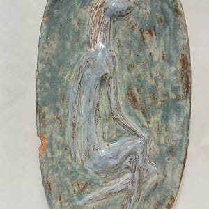May include: An oval ceramic wall plaque featuring a raised, stylized figure in a seated position. The piece has a glossy, mottled glaze in shades of blue, gray, and brown, with a textured surface and a small chip visible on the left edge.