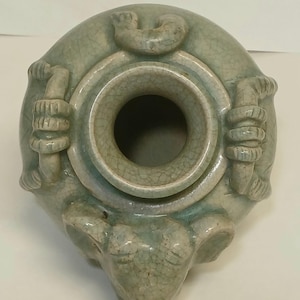 Puede incluir: Una vasija de cer&aacute;mica en color verde celad&oacute;n p&aacute;lido con un acabado de esmalte craquelado. El recipiente tiene una abertura circular en la parte superior, rodeada por tres asas esculpidas y un detalle de cabeza de animal en el cuerpo.