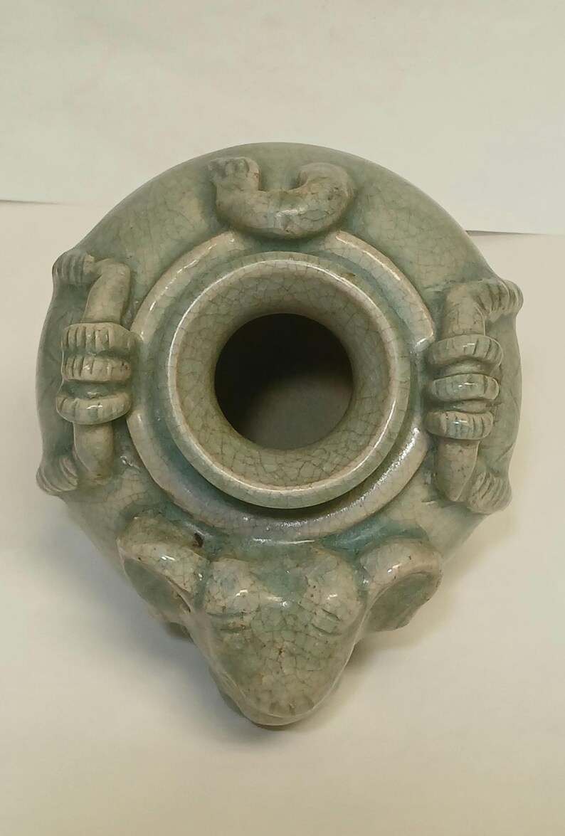 Puede incluir: Una vasija de cer&aacute;mica en color verde celad&oacute;n p&aacute;lido con un acabado de esmalte craquelado. El recipiente tiene una abertura circular en la parte superior, rodeada por tres asas esculpidas y un detalle de cabeza de animal en el cuerpo.
