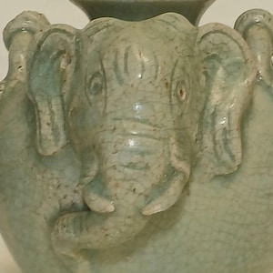 Puede incluir: Un jarr&oacute;n de cer&aacute;mica verde claro con un relieve prominente de cabeza de elefante en la parte frontal. La superficie del jarr&oacute;n tiene un acabado de esmalte craquelado visible, con la trompa, las orejas y los ojos del elefante bien definidos.