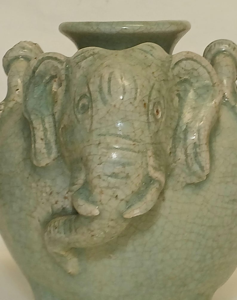 Puede incluir: Un jarr&oacute;n de cer&aacute;mica verde claro con un relieve prominente de cabeza de elefante en la parte frontal. La superficie del jarr&oacute;n tiene un acabado de esmalte craquelado visible, con la trompa, las orejas y los ojos del elefante bien definidos.