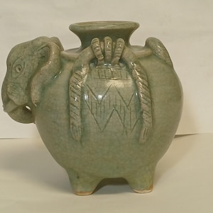 Puede incluir: Un recipiente de cer&aacute;mica de color verde claro con forma de elefante, cuerpo redondeado, una abertura circular central y tres patas cortas. La cabeza del elefante presenta ojos y orejas tallados, mientras que el cuerpo muestra patrones decorativos texturizados similares a cuerdas.