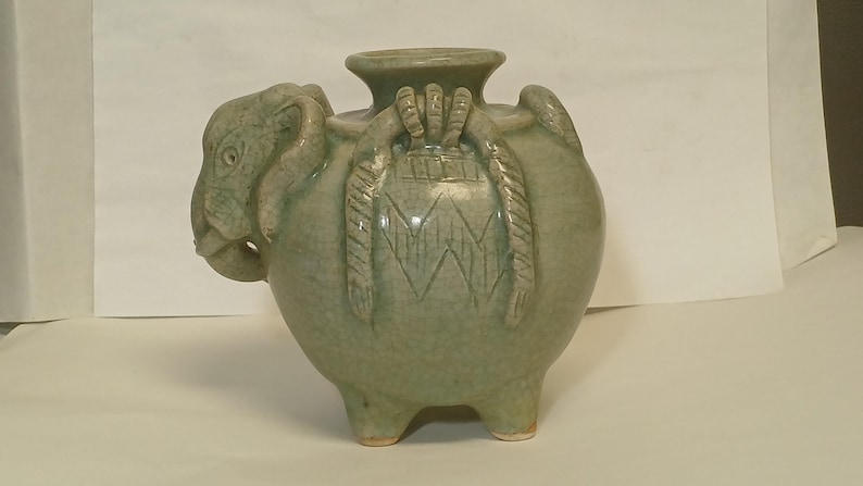 Puede incluir: Un recipiente de cer&aacute;mica de color verde claro con forma de elefante, cuerpo redondeado, una abertura circular central y tres patas cortas. La cabeza del elefante presenta ojos y orejas tallados, mientras que el cuerpo muestra patrones decorativos texturizados similares a cuerdas.