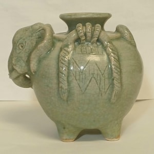 Puede incluir: Un recipiente de cer&aacute;mica verde claro con forma de elefante y acabado de esmalte craquelado. La pieza presenta una amplia abertura circular en la parte superior, tres patas cortas y patrones decorativos en relieve similares a cuerdas.
