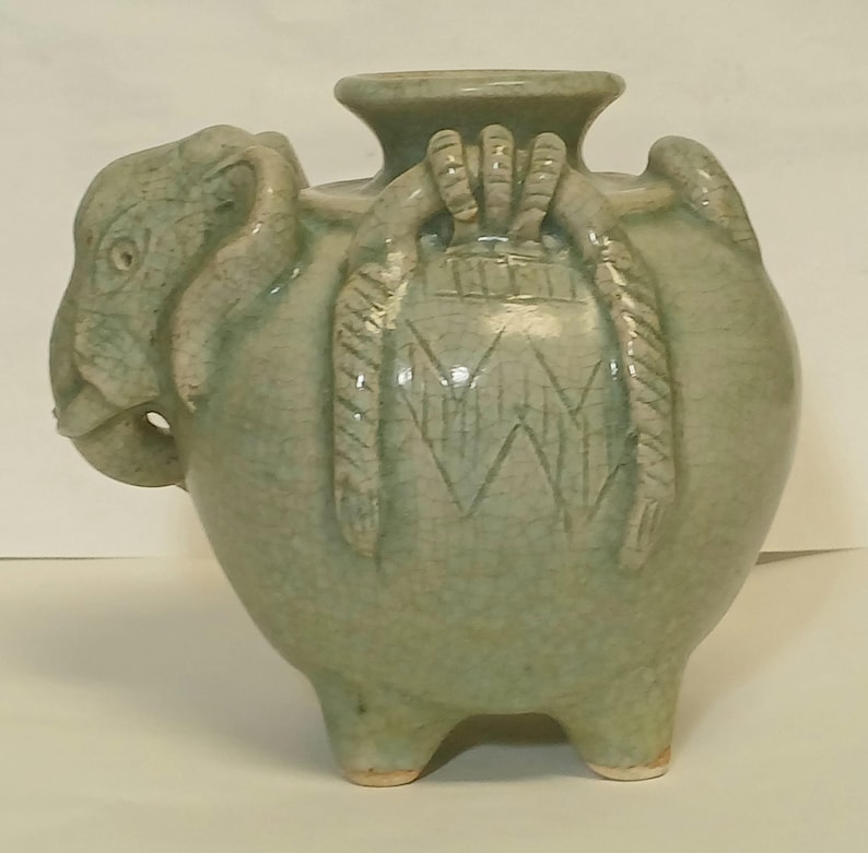 Puede incluir: Un recipiente de cer&aacute;mica verde claro con forma de elefante y acabado de esmalte craquelado. La pieza presenta una amplia abertura circular en la parte superior, tres patas cortas y patrones decorativos en relieve similares a cuerdas.
