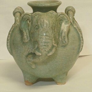 Puede incluir: Un jarr&oacute;n de cer&aacute;mica verde p&aacute;lido con acabado de esmalte craquelado, que presenta una cabeza de elefante esculpida en el frente y dos asas en los lados. El recipiente tiene un cuerpo ancho y redondeado, un borde circular ensanchado y tres patas.