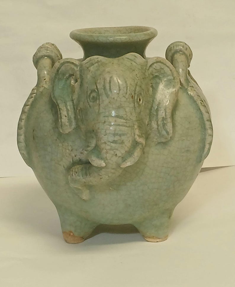 Puede incluir: Un jarr&oacute;n de cer&aacute;mica verde p&aacute;lido con acabado de esmalte craquelado, que presenta una cabeza de elefante esculpida en el frente y dos asas en los lados. El recipiente tiene un cuerpo ancho y redondeado, un borde circular ensanchado y tres patas.