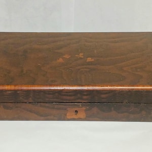 May include: A rectangular wooden box with a dark brown wood grain finish. The lid features a small, light-colored inlaid floral design in the center. The front panel includes a central keyhole escutcheon made of a lighter wood inlay.