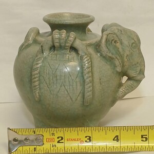 Puede incluir: Un jarr&oacute;n de cer&aacute;mica verde claro con acabado de esmalte craquelado, que presenta una cabeza de elefante esculpida en el lateral y asas decorativas texturizadas. Una cinta m&eacute;trica amarilla Stanley colocada delante muestra 10,16 cm.