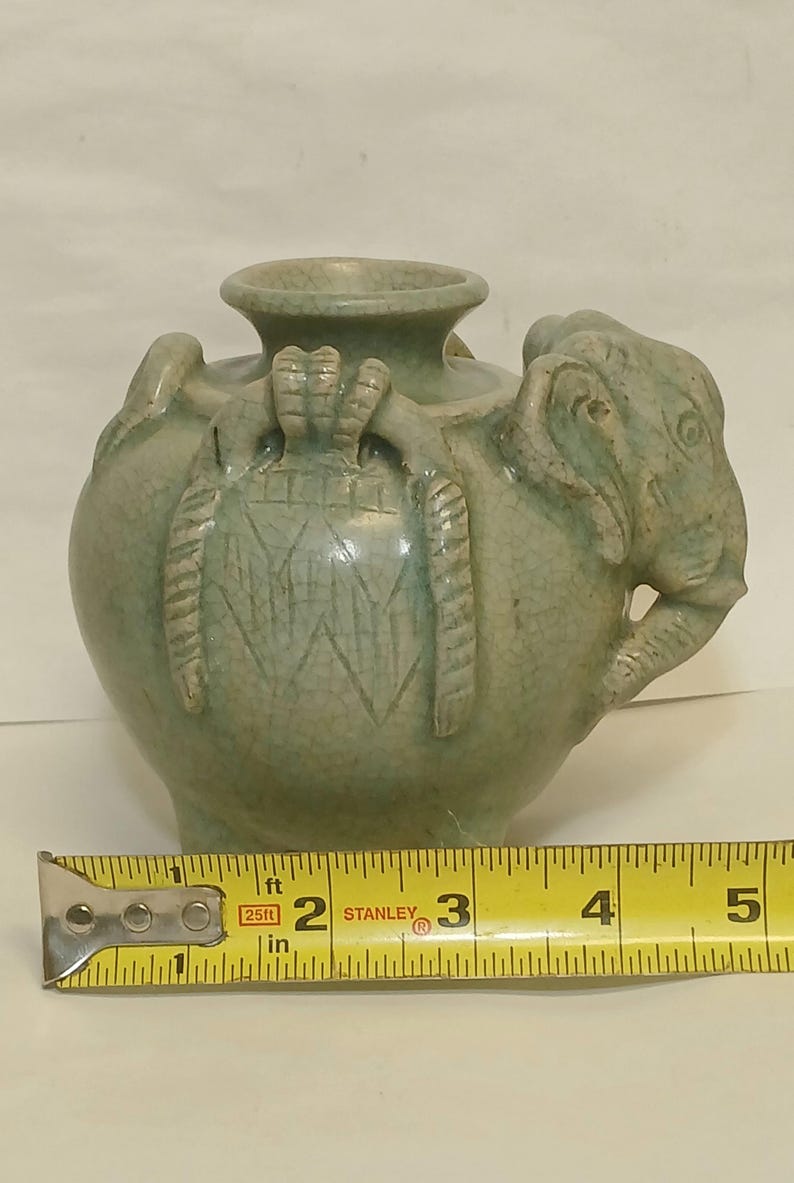 Puede incluir: Un jarr&oacute;n de cer&aacute;mica verde claro con acabado de esmalte craquelado, que presenta una cabeza de elefante esculpida en el lateral y asas decorativas texturizadas. Una cinta m&eacute;trica amarilla Stanley colocada delante muestra 10,16 cm.
