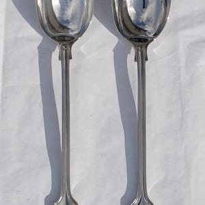 May include: A pair of polished silver-tone serving utensils consisting of a large solid spoon and a three-pronged serving fork. Both pieces feature long, slender handles with a decorative shell motif embossed at the base of each handle.