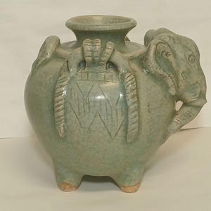 Puede incluir: Un recipiente de cerámica verde claro con acabado de esmalte craquelado, que presenta tres patas cortas y una escultura de cabeza de elefante integrada en el lateral. El cuerpo está decorado con patrones geométricos tallados y líneas.