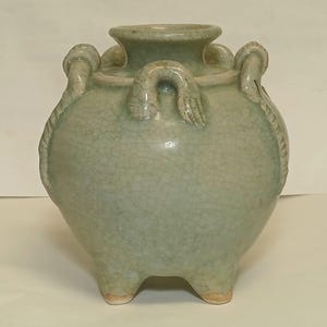 Puede incluir: Una vasija de cer&aacute;mica en color verde celad&oacute;n p&aacute;lido con un acabado de esmalte craquelado. El recipiente presenta un cuerpo ancho y redondeado, un cuello corto con borde ensanchado y tres pies peque&ntilde;os en la base. Tres asas curvas decorativas.