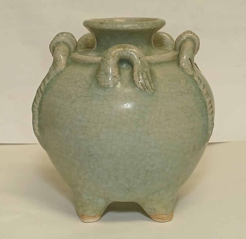 Puede incluir: Una vasija de cer&aacute;mica en color verde celad&oacute;n p&aacute;lido con un acabado de esmalte craquelado. El recipiente presenta un cuerpo ancho y redondeado, un cuello corto con borde ensanchado y tres pies peque&ntilde;os en la base. Tres asas curvas decorativas.