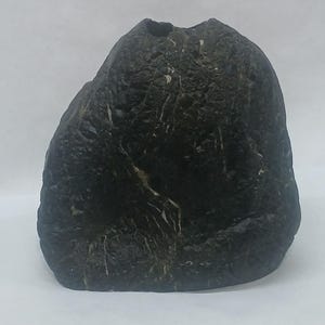 May include: A dark charcoal-colored vase with a rough, uneven, and textured surface. The vase has a rounded, organic shape with a small opening at the top. The exterior features subtle, lighter-colored veining patterns running through the dark stone-like material.
