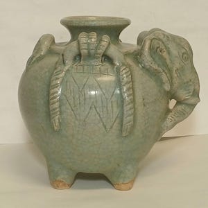 Puede incluir: Un recipiente de cer&aacute;mica celad&oacute;n verde claro con acabado de esmalte craquelado. El cuerpo redondo presenta tres pies cortos, un cuello ancho y una cabeza de elefante esculpida en el lateral. La superficie est&aacute; decorada con patrones geom&eacute;tricos.