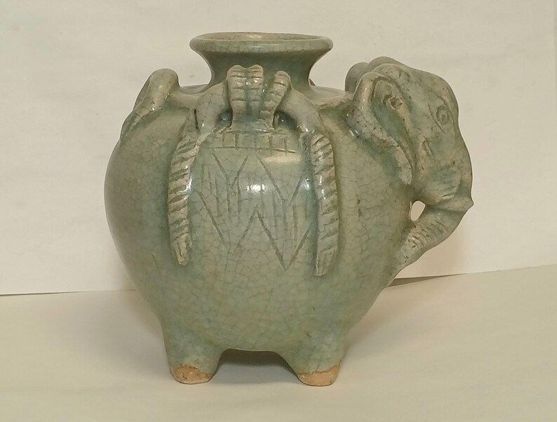 Puede incluir: Un recipiente de cer&aacute;mica celad&oacute;n verde claro con acabado de esmalte craquelado. El cuerpo redondo presenta tres pies cortos, un cuello ancho y una cabeza de elefante esculpida en el lateral. La superficie est&aacute; decorada con patrones geom&eacute;tricos.