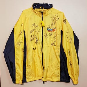May include: A bright yellow and navy blue windbreaker jacket featuring a full-length front zipper, a high collar, and long sleeves. The jacket is covered in numerous black ink signatures and displays the O'Reilly Racing logo on the chest.