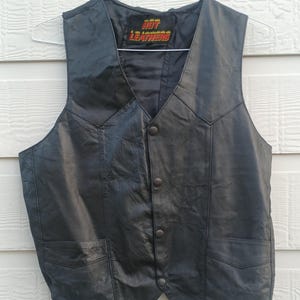 May include: A black leather vest featuring a V-neckline, four front snap button closures, and two lower pockets. The interior label displays the text Hot Leathers in red and yellow lettering. The garment is sleeveless with a pointed hemline.
