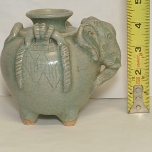 Puede incluir: Un recipiente de cer&aacute;mica verde claro con forma de elefante y acabado de esmalte craquelado, que presenta una abertura central y tres patas peque&ntilde;as. Una cinta m&eacute;trica amarilla est&aacute; junto al objeto, mostrando una altura de 12,7 cm.