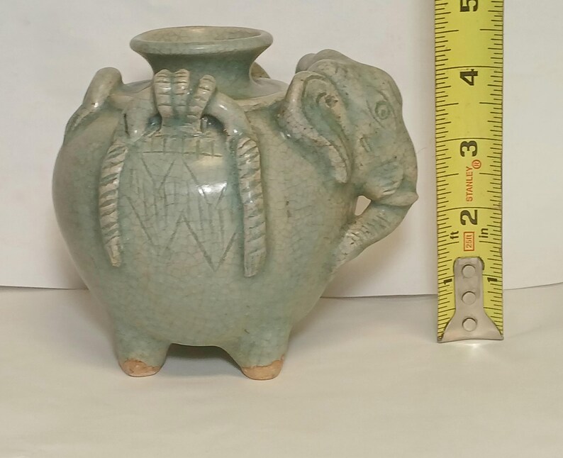 Puede incluir: Un recipiente de cer&aacute;mica verde claro con forma de elefante y acabado de esmalte craquelado, que presenta una abertura central y tres patas peque&ntilde;as. Una cinta m&eacute;trica amarilla est&aacute; junto al objeto, mostrando una altura de 12,7 cm.