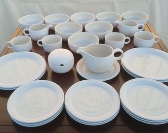 Op de afbeelding: Een set wit keramieken servies bestaande uit meerdere borden, kommen, kopjes en kannetjes. Het servies heeft een gladde, effen witte afwerking met subtiele cirkelvormige ribbels aan de buitenkant van de kopjes en de suikerpot met deksel.