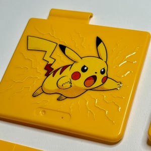 May include: A yellow plastic case featuring a Pikachu graphic. The cartoon character is depicted with red cheeks and a lightning bolt tail. The case has a textured surface and a small tab at the top.