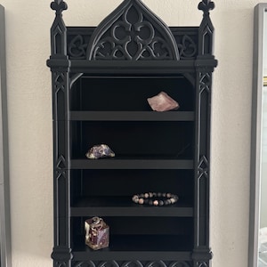 May include: Black gothic-style wall shelf with four shelves. The shelf is decorated with ornate details and pointed arches. The shelves display a pink crystal, a purple geode, a beaded bracelet, and a small decorative cube.