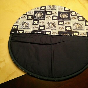 May include: A round fabric tortilla warmer featuring a navy blue and light blue checkered pattern with the University of North Carolina Tar Heels logo and text printed repeatedly. The bottom half is solid navy blue fabric with a circular stitched edge.